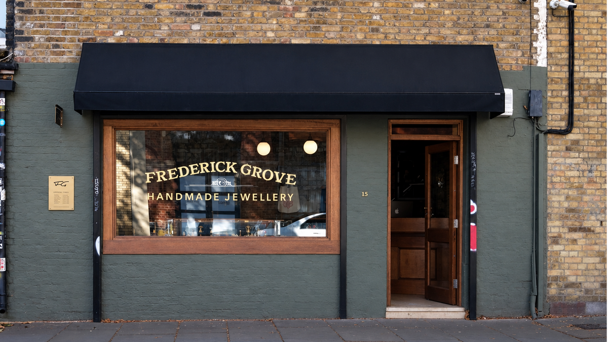 A storefront facade for Frederick Grove Handmade Jewellery, featuring a dark green painted wall, a black awning, a large window with gold lettering, and an open wooden door on a brick building.